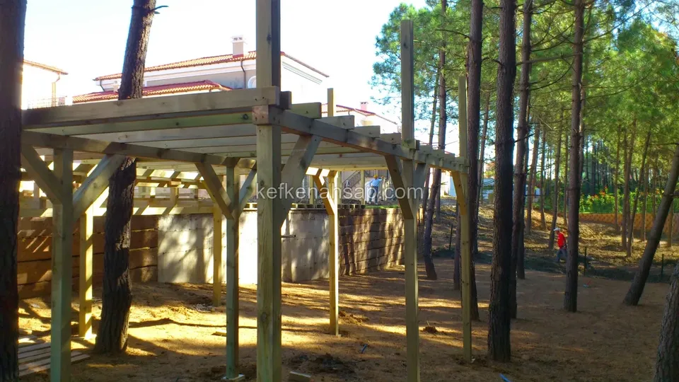 Çam ağaçları arasında eğimli arazide yapım aşamasındaki ahşap pergola iskeleti ve çapraz payandalar