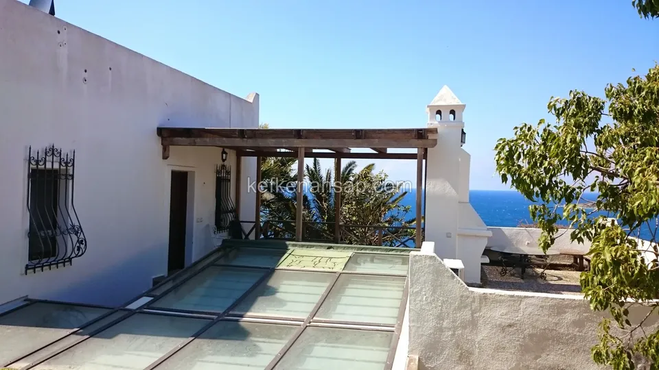 Beyaz evin terasında cam zeminin üzerinde ahşap pergola, arkada deniz ve bir ağaç