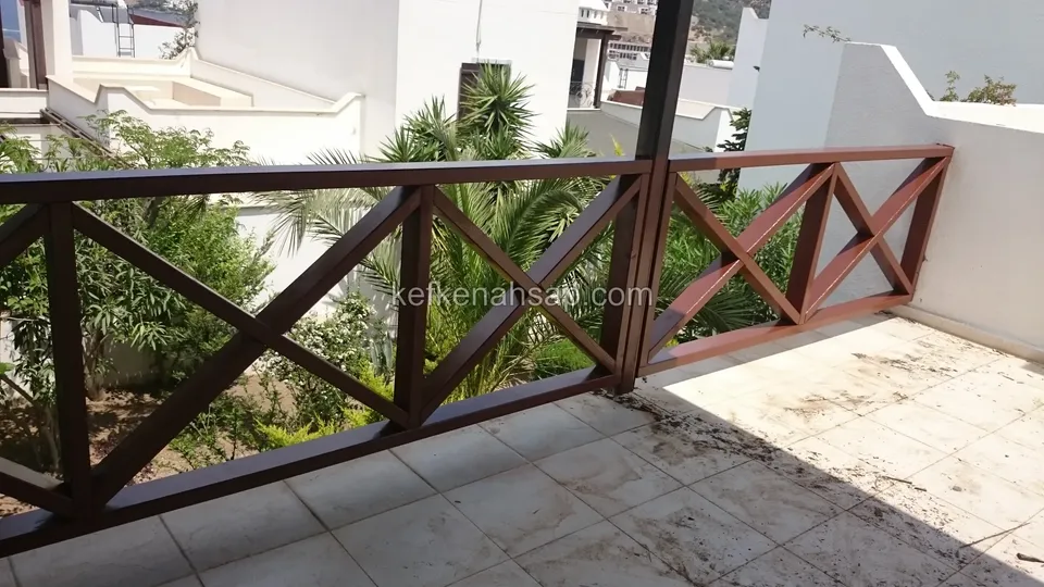 Dark brown varnished timber railing with cross braces on the edge of a tiled balcony, with a palm and white walls behind