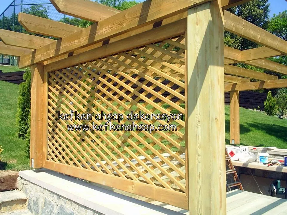 Bahçede kalın ahşap kolonlu pergola, çapraz kafes panel ve üstte yatay kirişler; arkada çim ve ağaçlar görünüyor