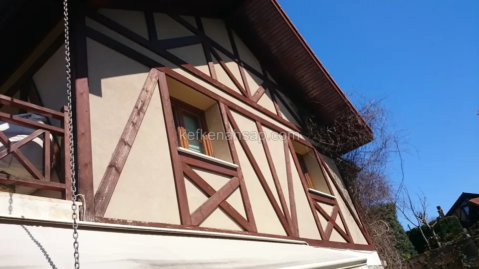 Under a blue sky, a facade with diagonal timber framing, small windows, a balcony railing and wide roof eaves