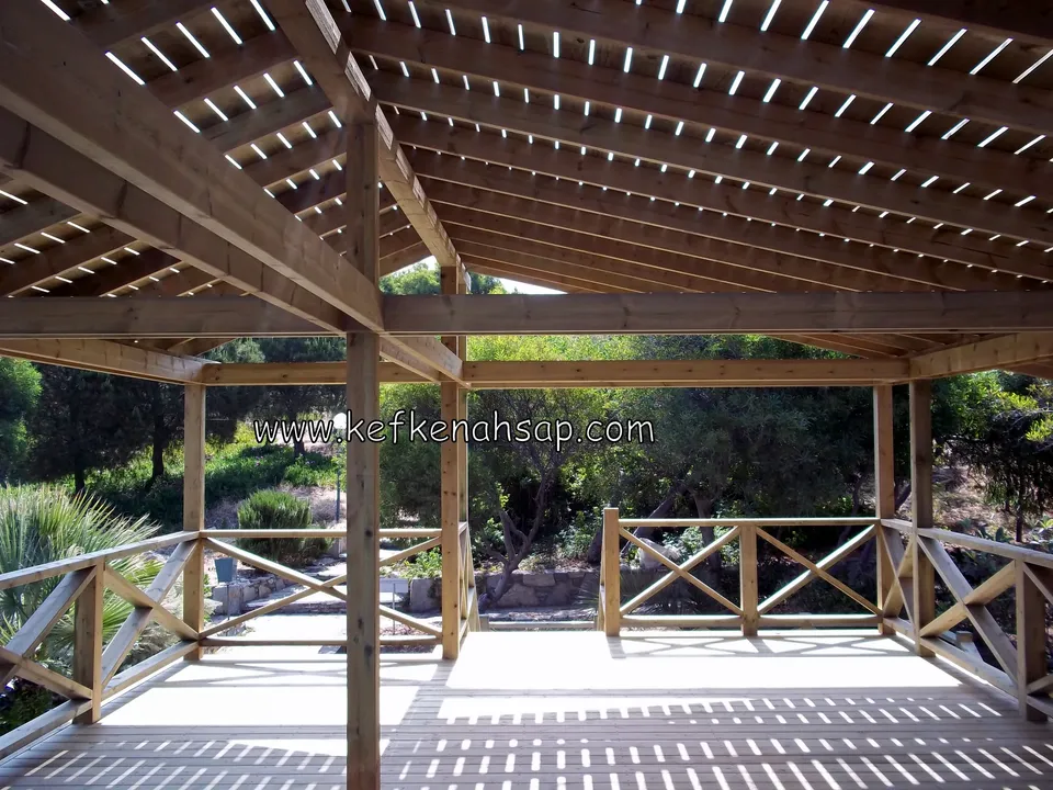 Çapraz korkuluklu açık renkli deck üzerinde pergola çatı kirişleri, arkada yeşil bitki örtüsü