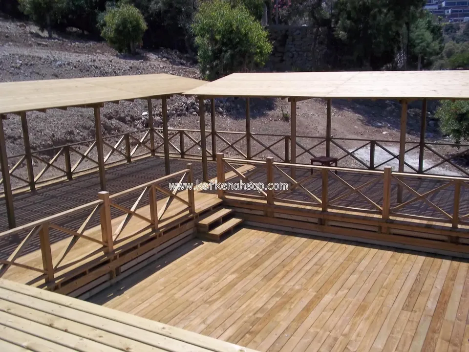 Eğimli bir arazide çapraz korkuluklu ahşap deck, basamaklar ve düz ahşap çatılı iki açık gölgelik