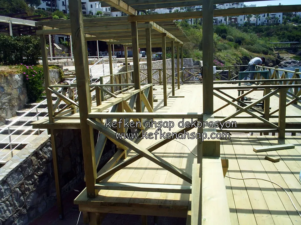 Taş duvarın yanında çapraz çıtalı korkuluklu deck ve üzerinde yapımı süren ahşap pergola iskeleti