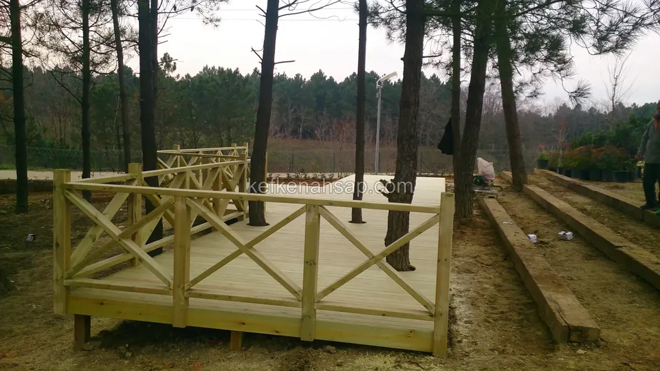 Helle Terrasse mit Kreuzstreben-Geländer zwischen Kiefern, daneben in Reihen in den Boden gesetzte Holzschwellen