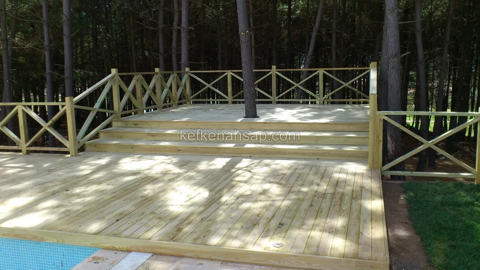 Çam ağaçları arasında, çapraz çıtalı korkuluklu geniş açık renkli deck; arkada basamaklar ve sol altta havuz kenarı