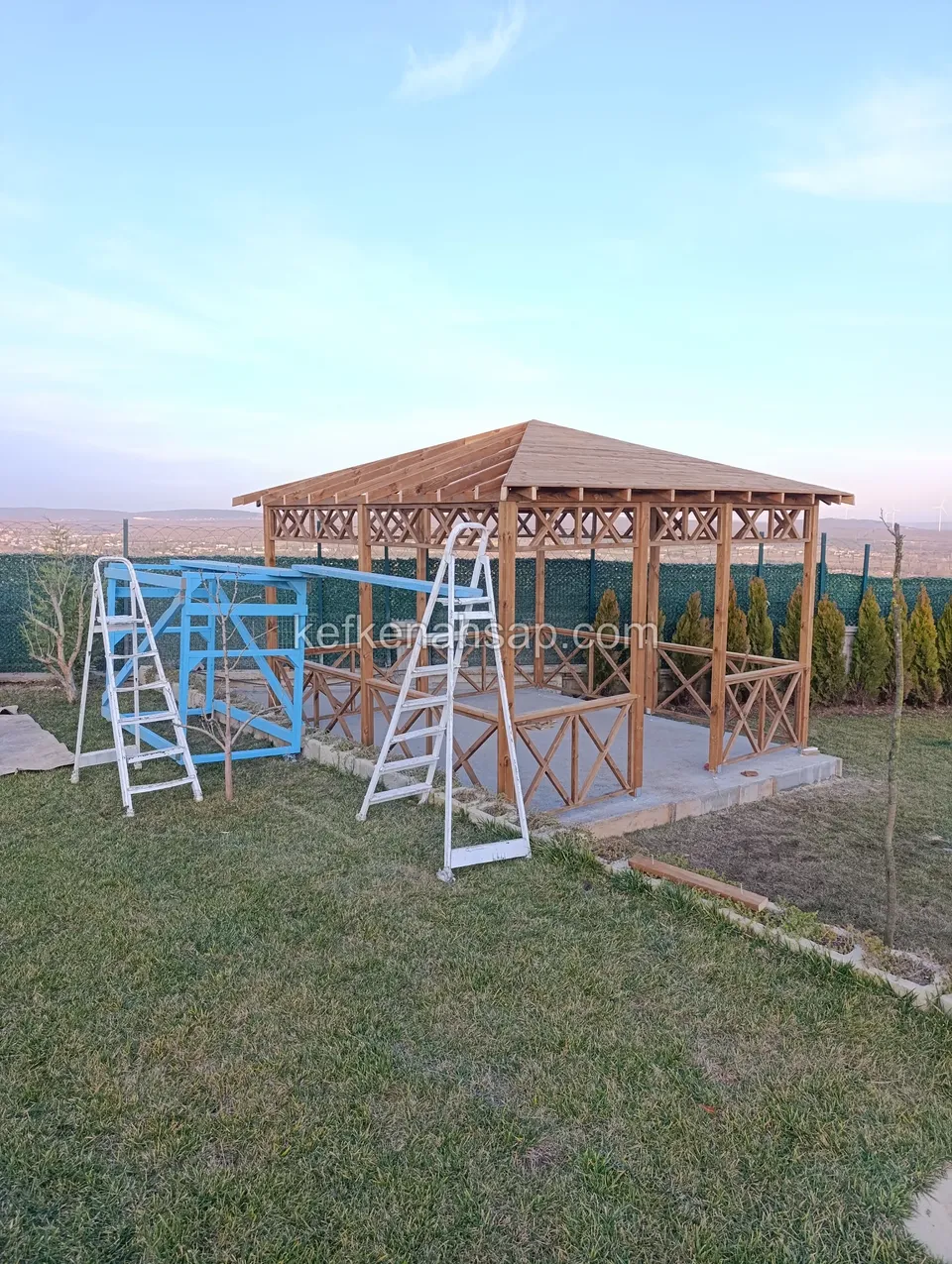 Çapraz korkuluklu kamelyanın önünde iki beyaz merdiven ve mavi iskele parçaları, çimlik bahçe