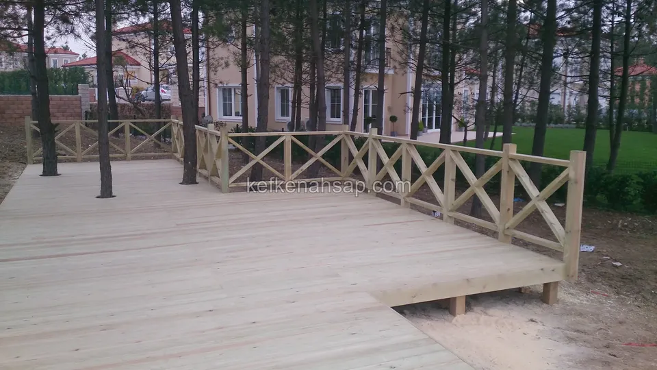 Çam ağaçları ve villaların önünde köşe yapan L şekilli açık renkli deck; X çıtalı korkuluk ve yan tarafta çıplak toprak