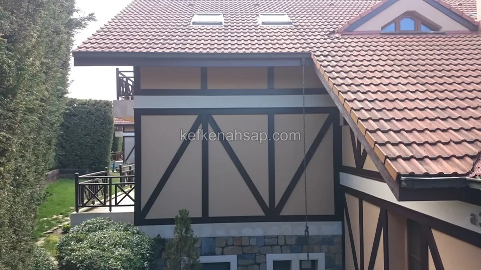 Krem sıvalı yüzeyde koyu ahşap çaprazlı kirişlerle bölünmüş cephe, üstte kiremit çatı ve solda ahşap korkuluklu balkon