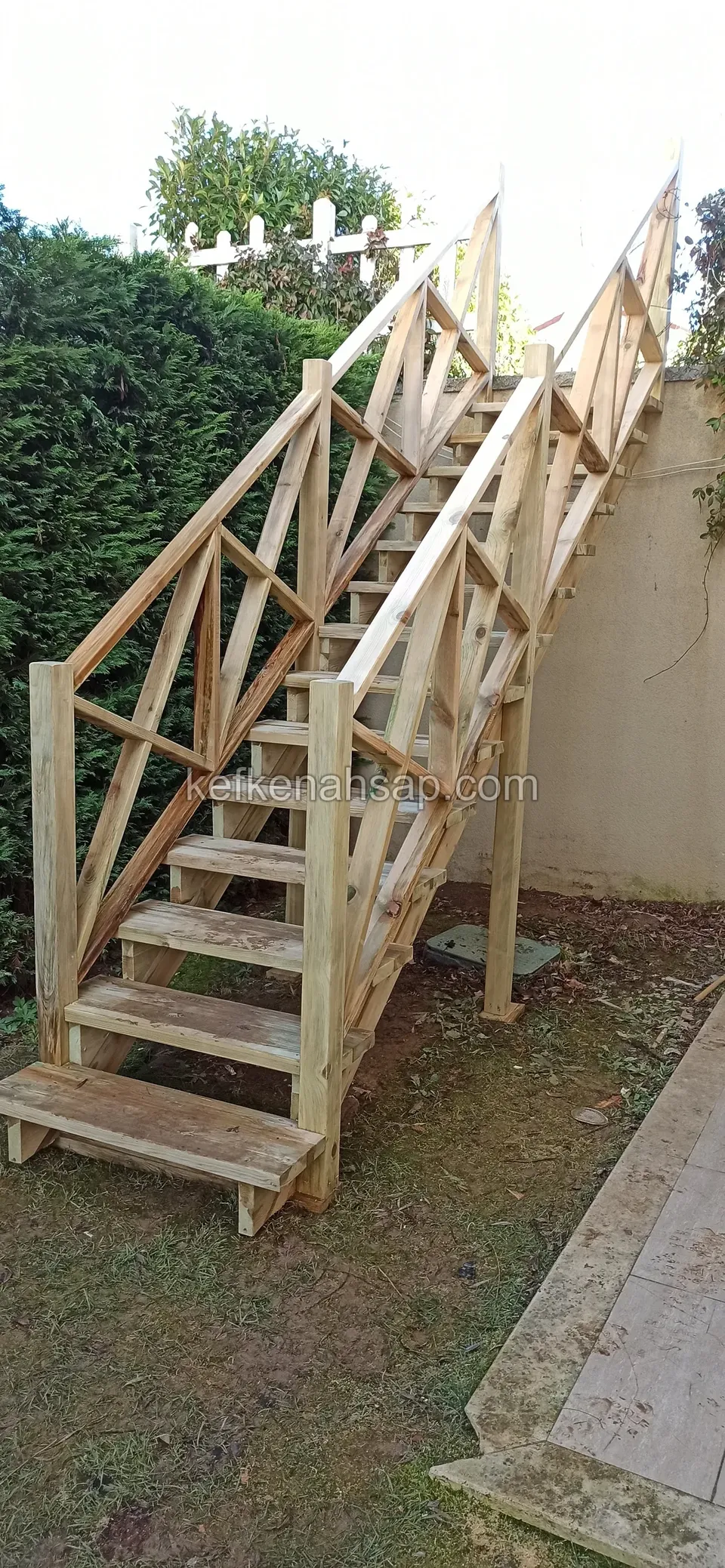 Holztreppe, an der das Kreuzstreben-Geländer montiert wird, am Boden ein Kabel, daneben grüne Hecke und Mauer