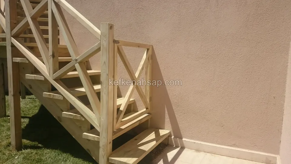Pembe boyalı duvarın önünde çaprazlı korkuluklu ahşap merdiven; ilk basamak çimde duruyor