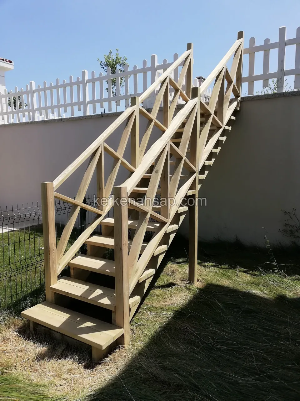 Holztreppe mit Kreuzstreben-Geländer, dahinter Mauer und weißer Lattenzaun, davor hohes Gras