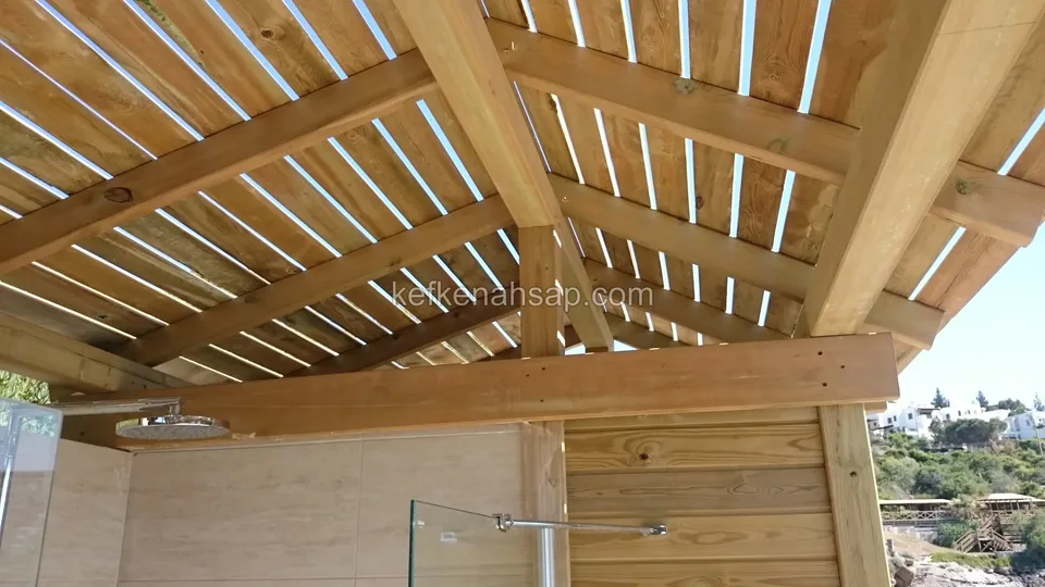 Ahşap çatı kirişlerinin altında cam bölmeli ve ahşap kaplı duş alanı, duvarda yağmurlama başlığı
