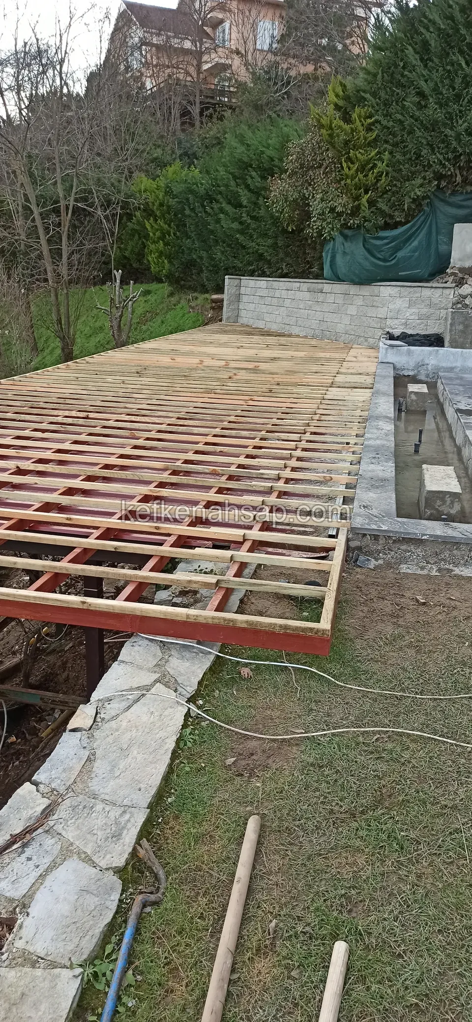 Çelik çerçeve üzerine ahşap çıtaların sıralandığı geniş deck alt konstrüksiyonu, yamaçta bir bahçe önünde