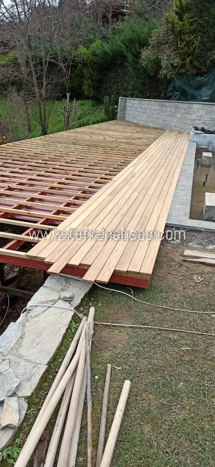 Bahçede kırmızı boyalı çelik iskelet üzerine döşenmeye başlanmış ahşap deck tahtaları, yanında taş duvar ve havuz boşluğu
