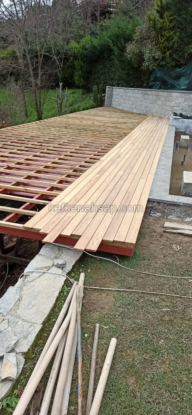 Bahçede kırmızı çelik iskelet üzerine döşenen ahşap deck; bir kısmı tahtalı, bir kısmı alt konstrüksiyon açıkta