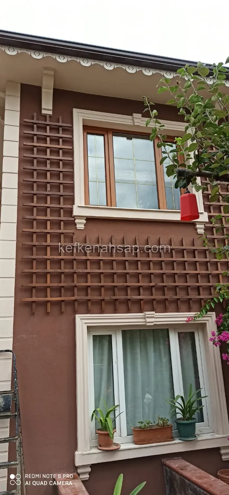 Kahverengi boyalı ev cephesinde iki pencere arasına monte edilmiş ahşap çıta ızgara, yanında çiçekli sarmaşık bitkiler