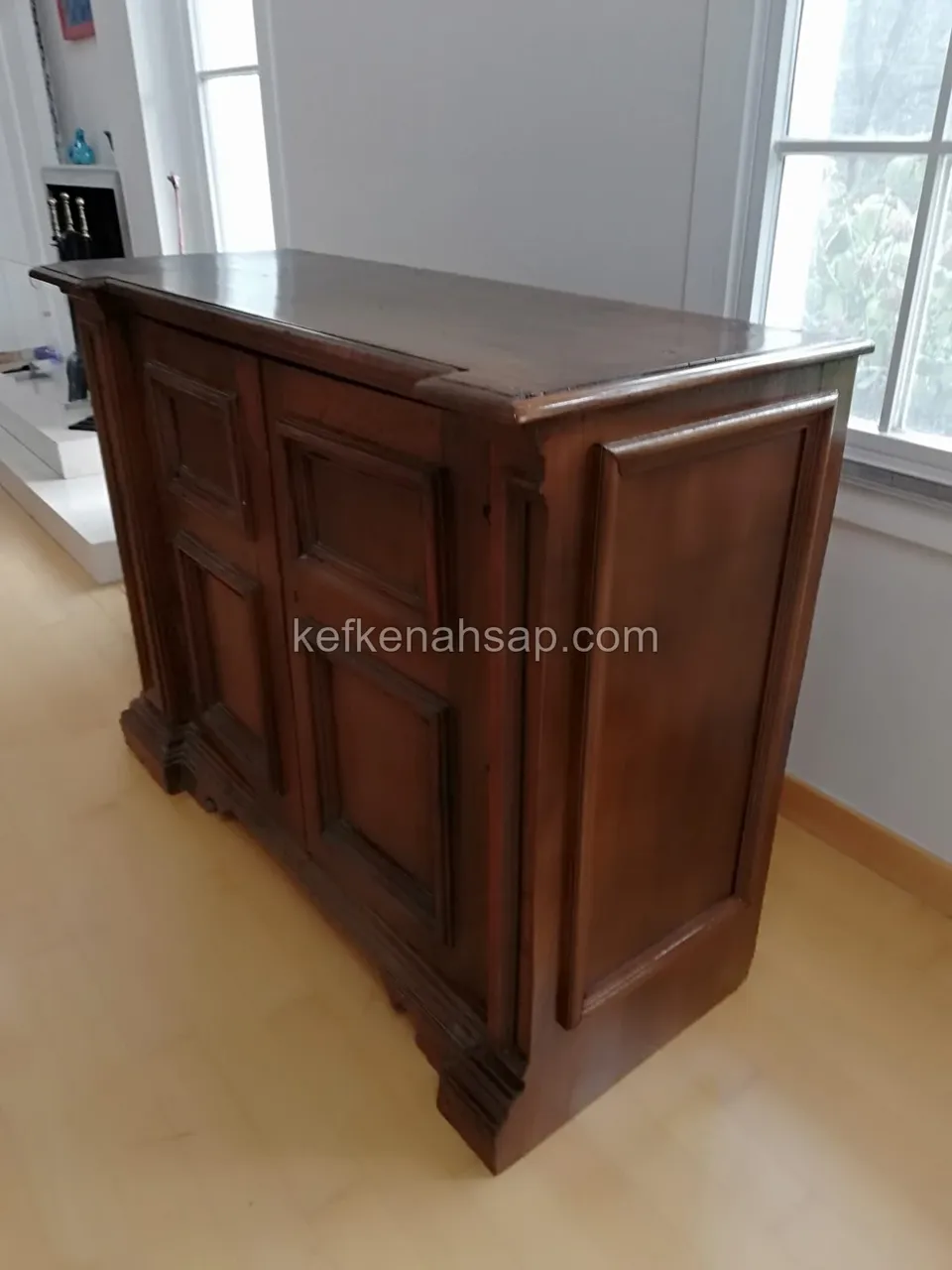 Pencere önünde duran, çerçeveli kapakları ve parlak cilalı üstü olan koyu kahverengi ahşap dolap