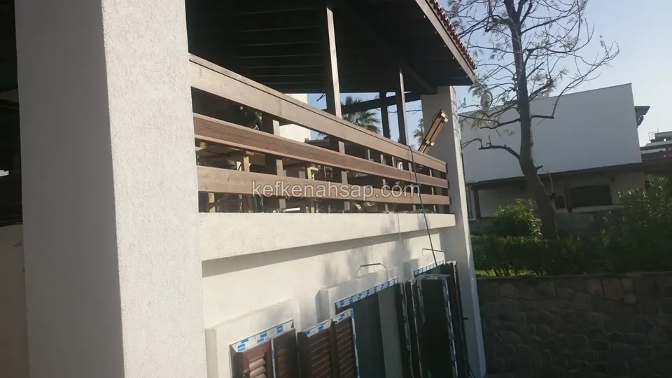 Çift katlı evin üst terasında yatay çıtalı ahşap korkuluk ve çatı; alt katta panjurlu pencereler