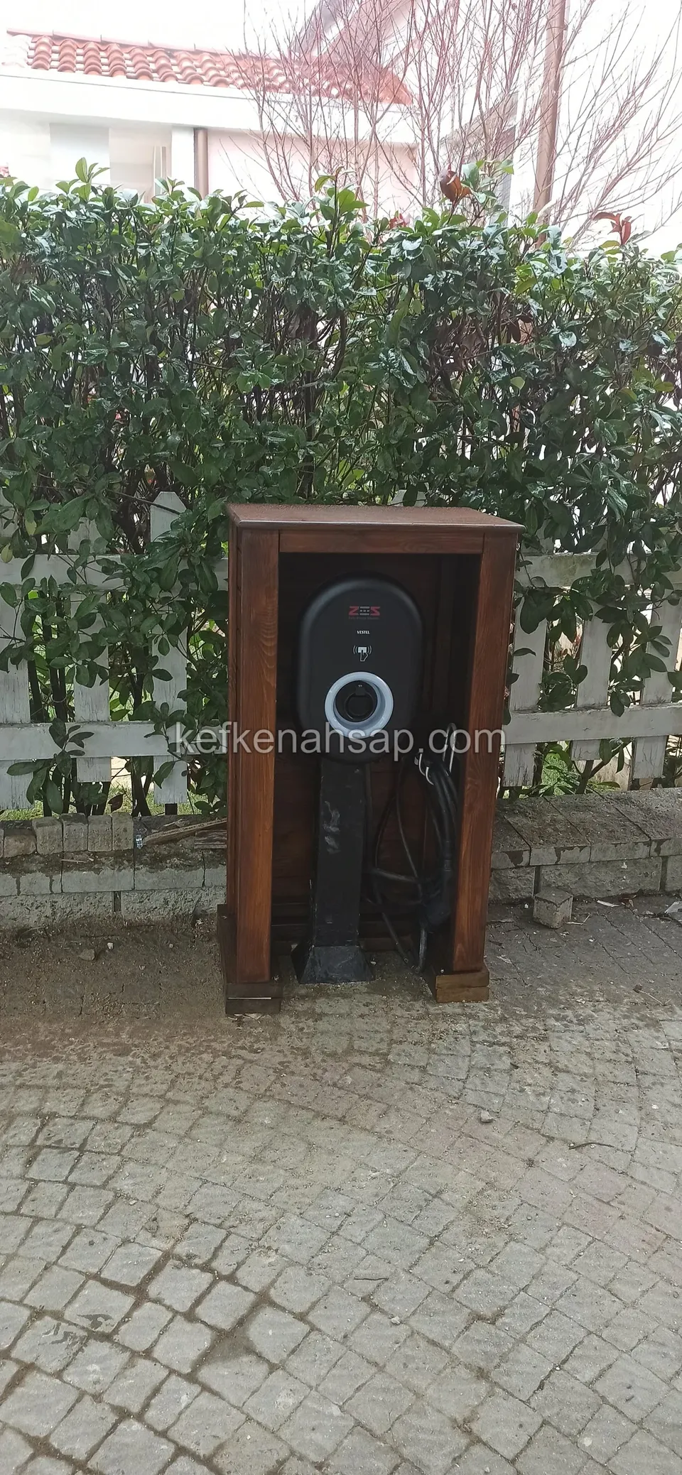 Taş döşemeli avluda, çalı çitinin önünde duran, ön yüzü açık ve içinde yuvarlak ışıklı bir cihaz bulunan koyu ahşap kabin