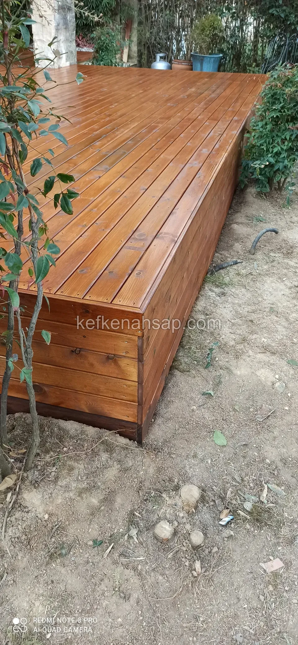 Glossy varnished orange-brown raised deck corner with side cladding and bare soil around it