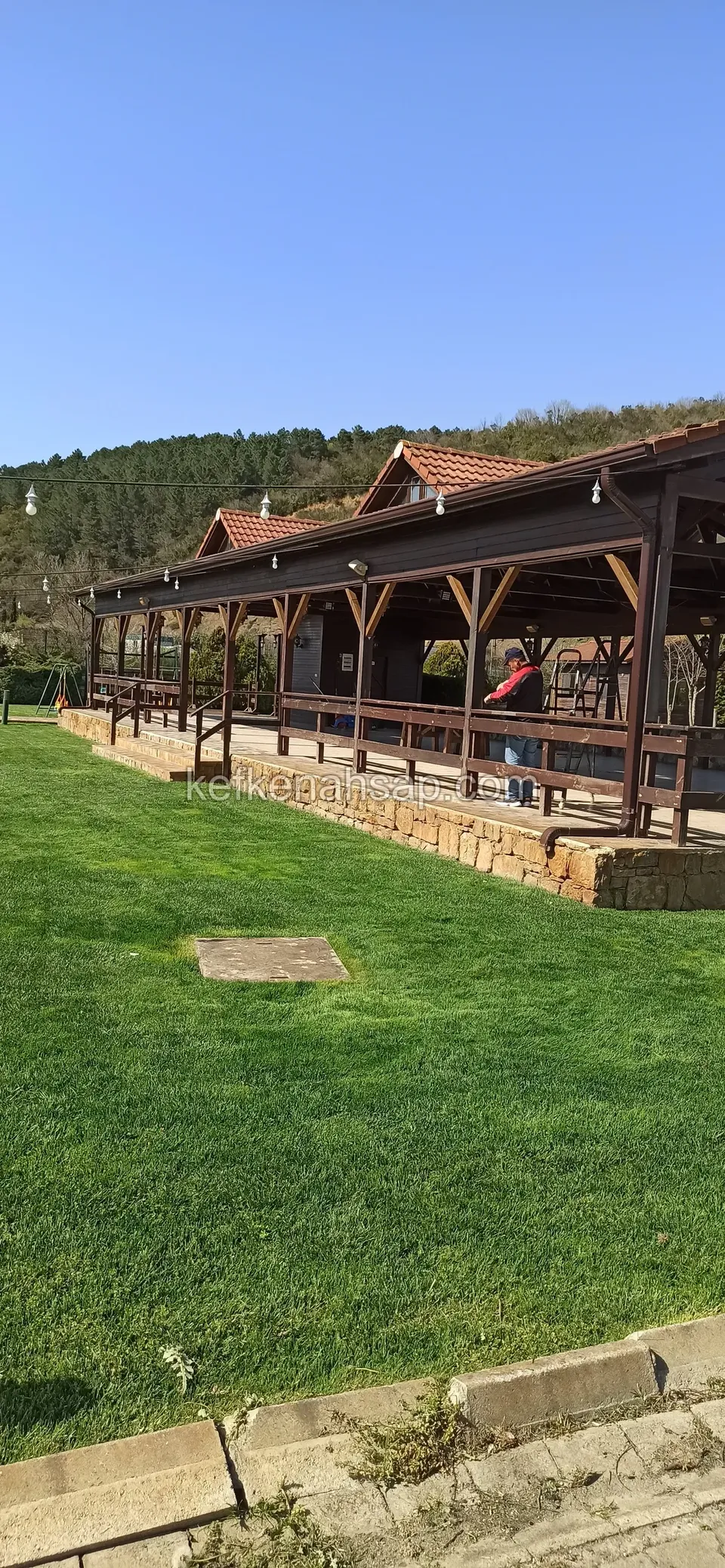Çim alanın kenarında duran, kiremit çatılı ve çevresi açık uzun ahşap yapı