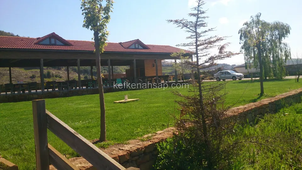 Geniş çim alanın karşısında duran, kırmızı kiremit çatılı ve yanları açık uzun ahşap yapı