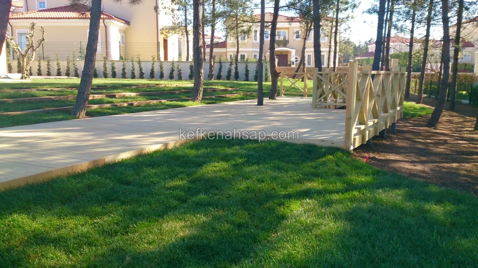 Çimlik alandan çam ağaçlarına doğru uzanan açık renkli deck yolu; ucunda çapraz çıtalı korkuluk