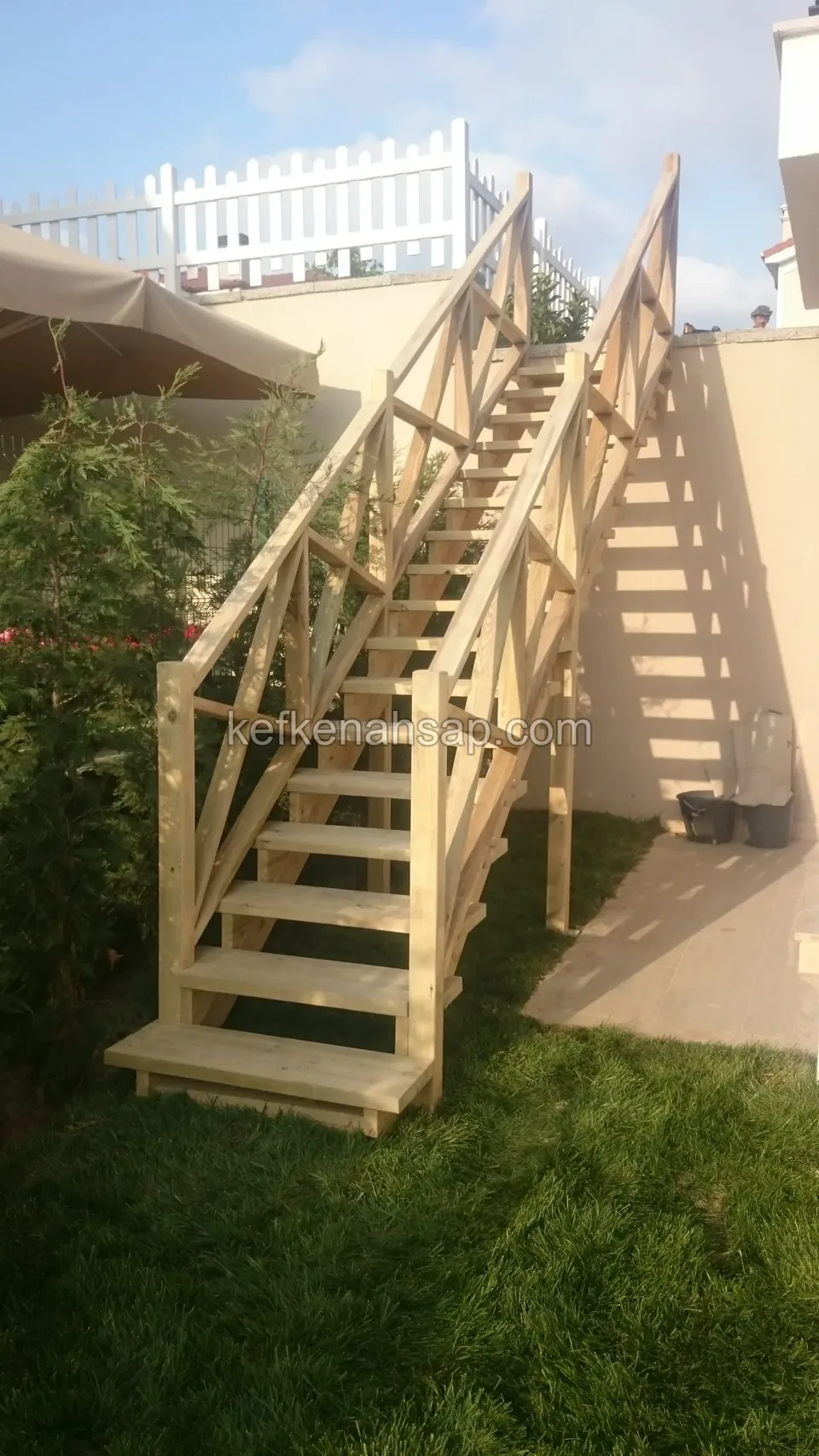 Holztreppe mit Kreuzstreben-Geländern vom Rasen zum Bereich über der Mauer, oben ein weißer Lattenzaun