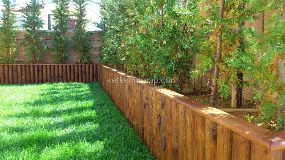 Low brown timber wall of vertical boards enclosing the lawn on three sides, with slim-trunked trees and shrubs behind