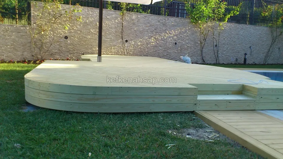 Große Holzplattform am Rasenrand, ihre Seiten aus gebogenen Brettern, dazu ein schmaler Steg zum Pool