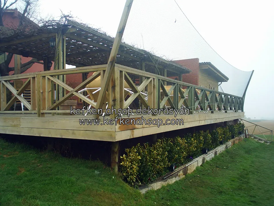 Çimenlik kenarında bir binanın önünde çapraz X korkuluklu, pergola çatılı yükseltilmiş deck, altında çalılar