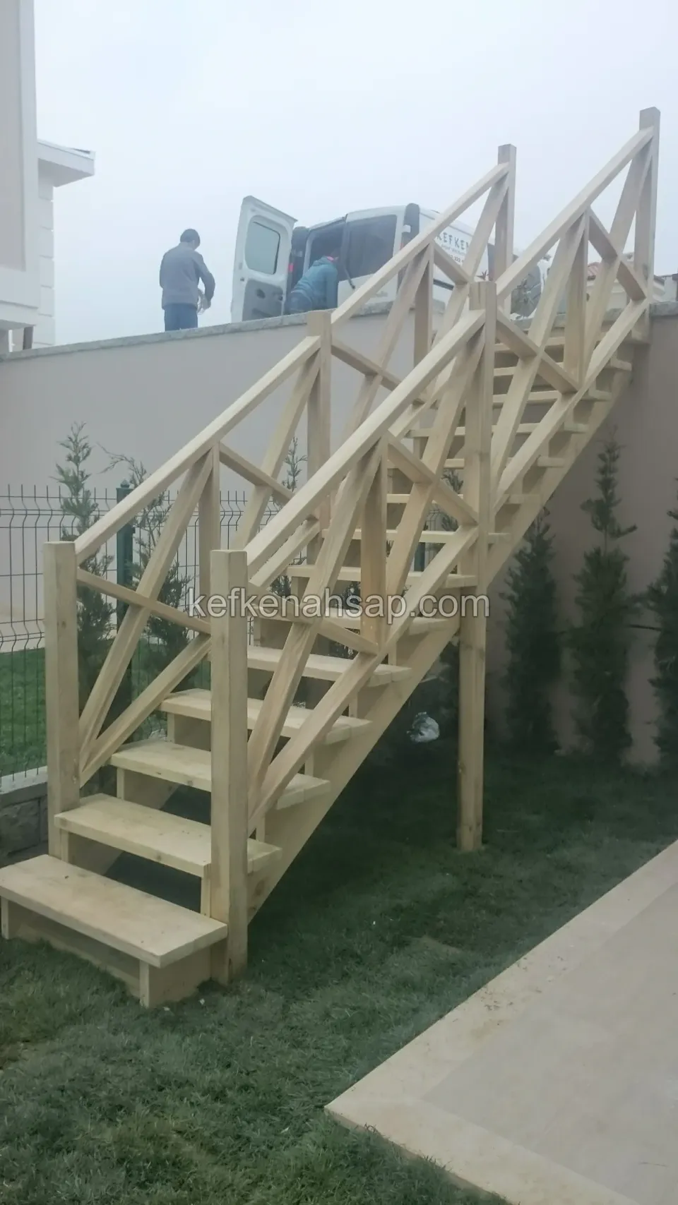 Yeni serilmiş çim üzerinde ayaklı ahşap merdiven ve çaprazlı korkuluk; arkada duvar ve sisli hava