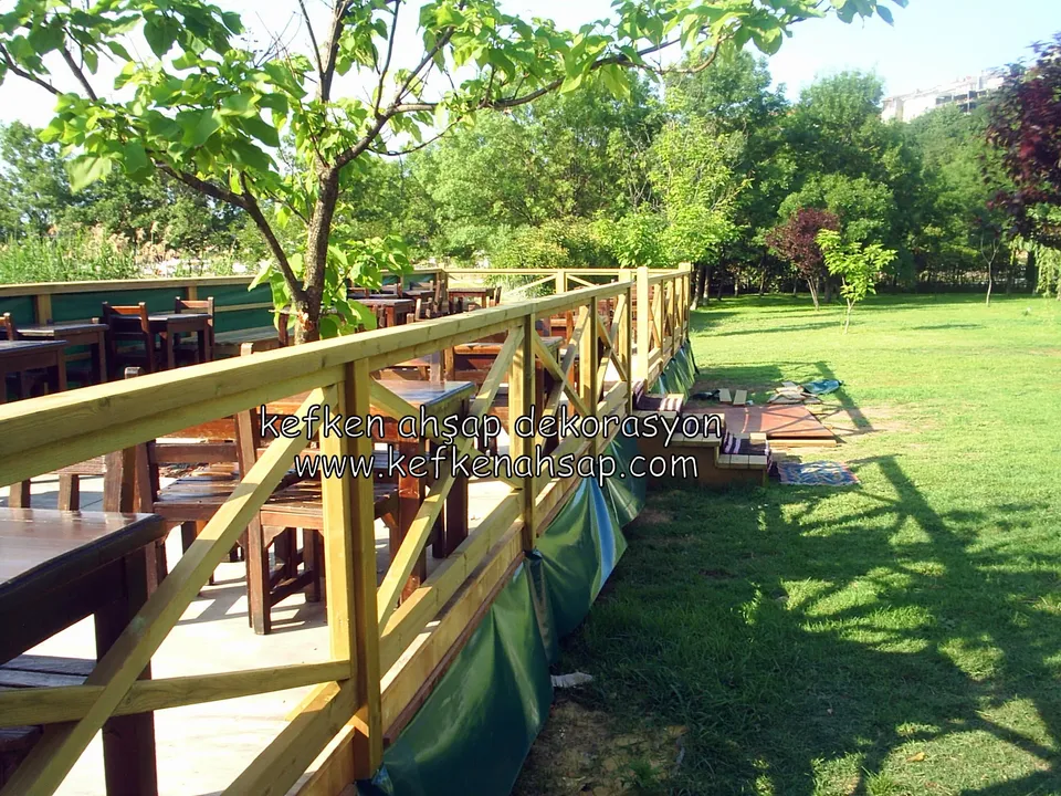 Çimlik kenarındaki deck terasın çevresinde sarımsı çapraz çıtalı ahşap korkuluk, içeride ahşap masa ve sandalyeler