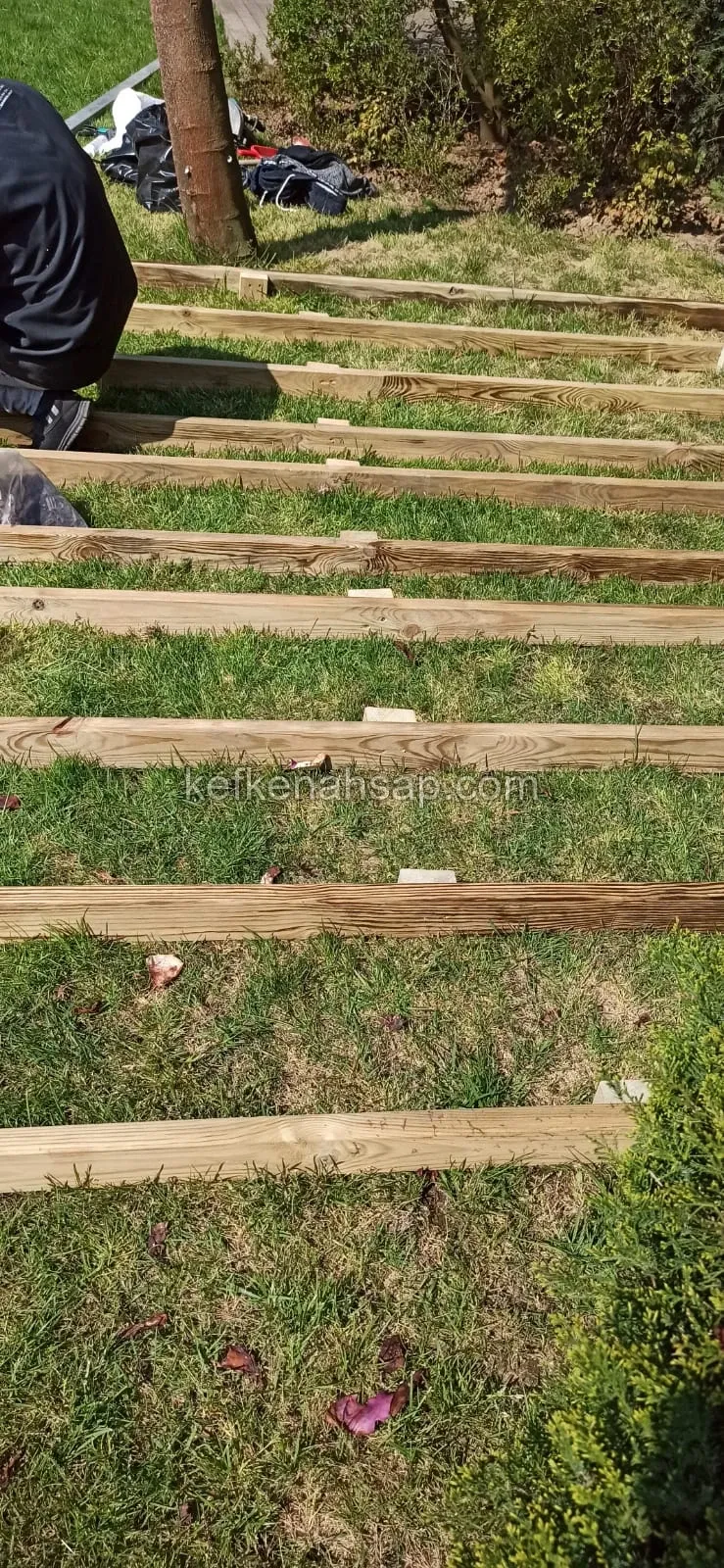 Çimlik zemine yan yana serilmiş ahşap taşıyıcı kirişler, kenarda çalışan bir usta ve arkada ağaç gövdesi