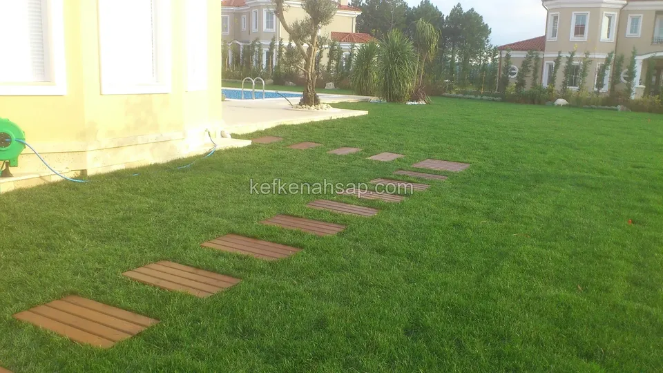 Geniş çimlikte diyagonal bir hat izleyen turuncu-kahve ahşap basamak plakaları; arkada havuz ve sarı cepheli villa