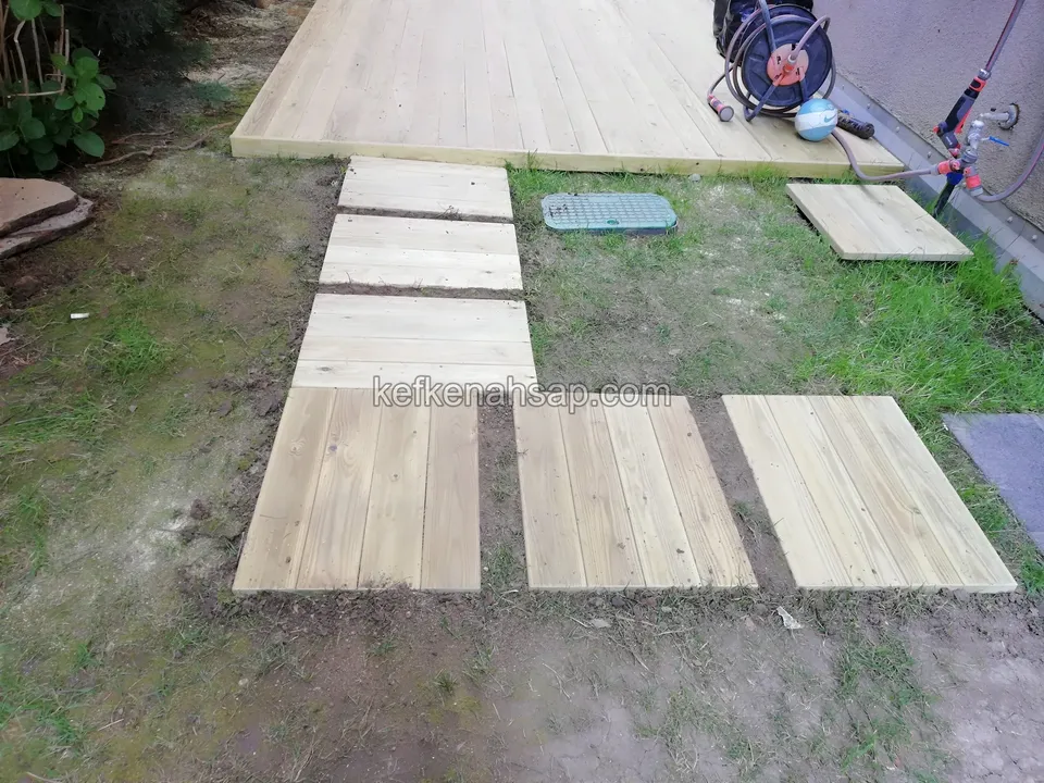 Toprak ve çimde sıralanmaya başlayan açık renk ahşap basamak plakaları; ucunda deck alanı, yerde kablo makarası