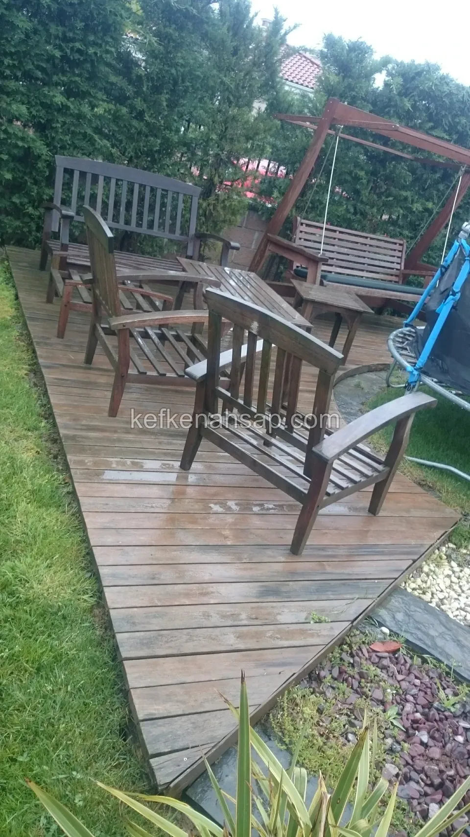 Auf einer sonnengrauen Deckplattform im Rasengarten stehen ein Holztisch, Sessel mit Armlehnen