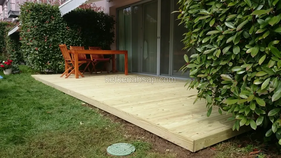 Çimenin kenarında, cam kapının önüne döşenmiş yeni açık renkli deck; üstünde turuncu ahşap bir masa ve sandalyeler