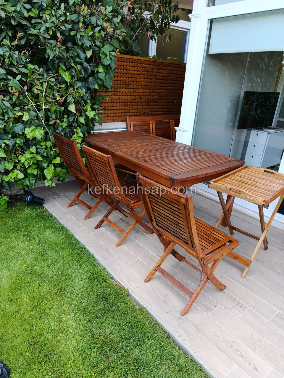At the edge where a pale tiled terrace meets the lawn, a long wooden dining table