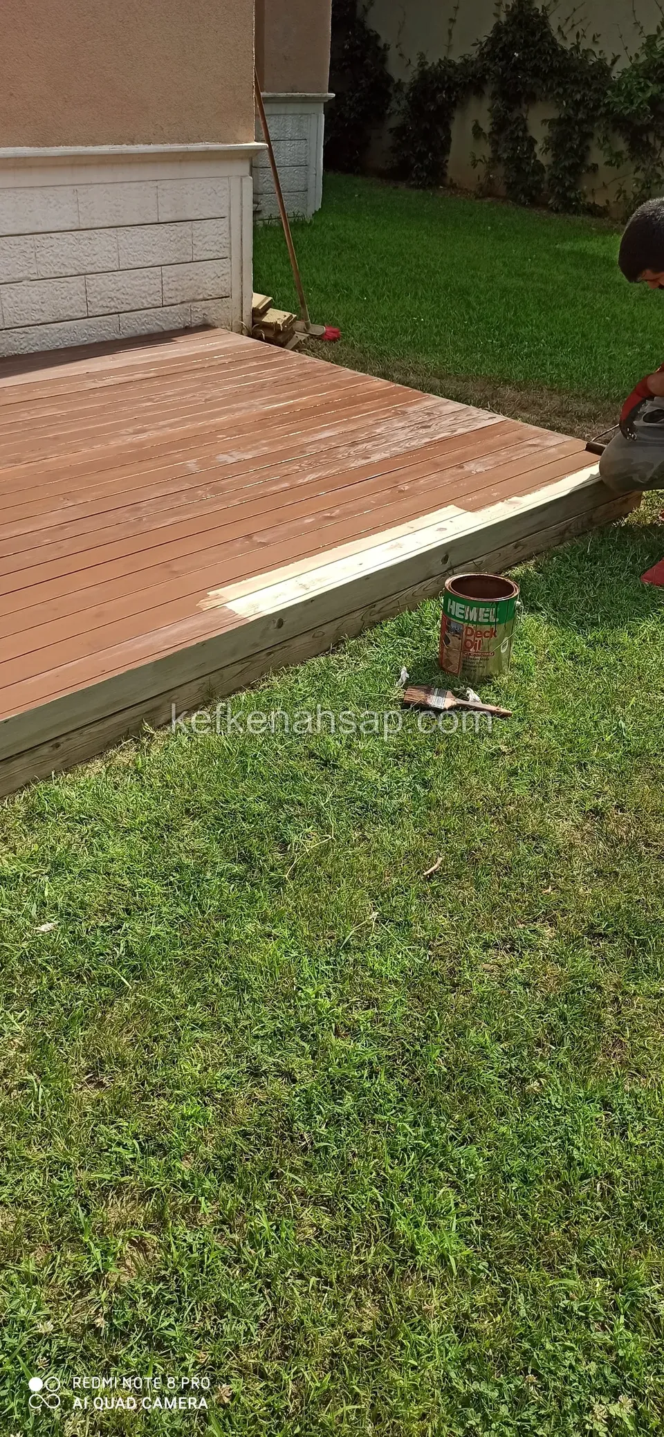 Deck, das am Rasenrand braun gestrichen wird, daneben Farbdose und Pinsel