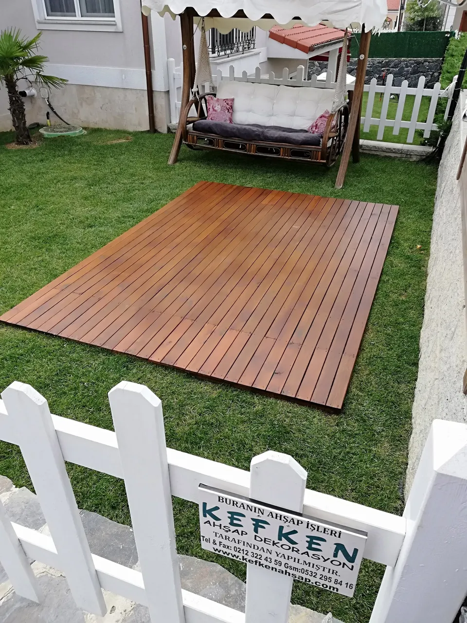 Brown rectangular deck laid on the lawn, with a cushioned garden swing behind and a white fence in front