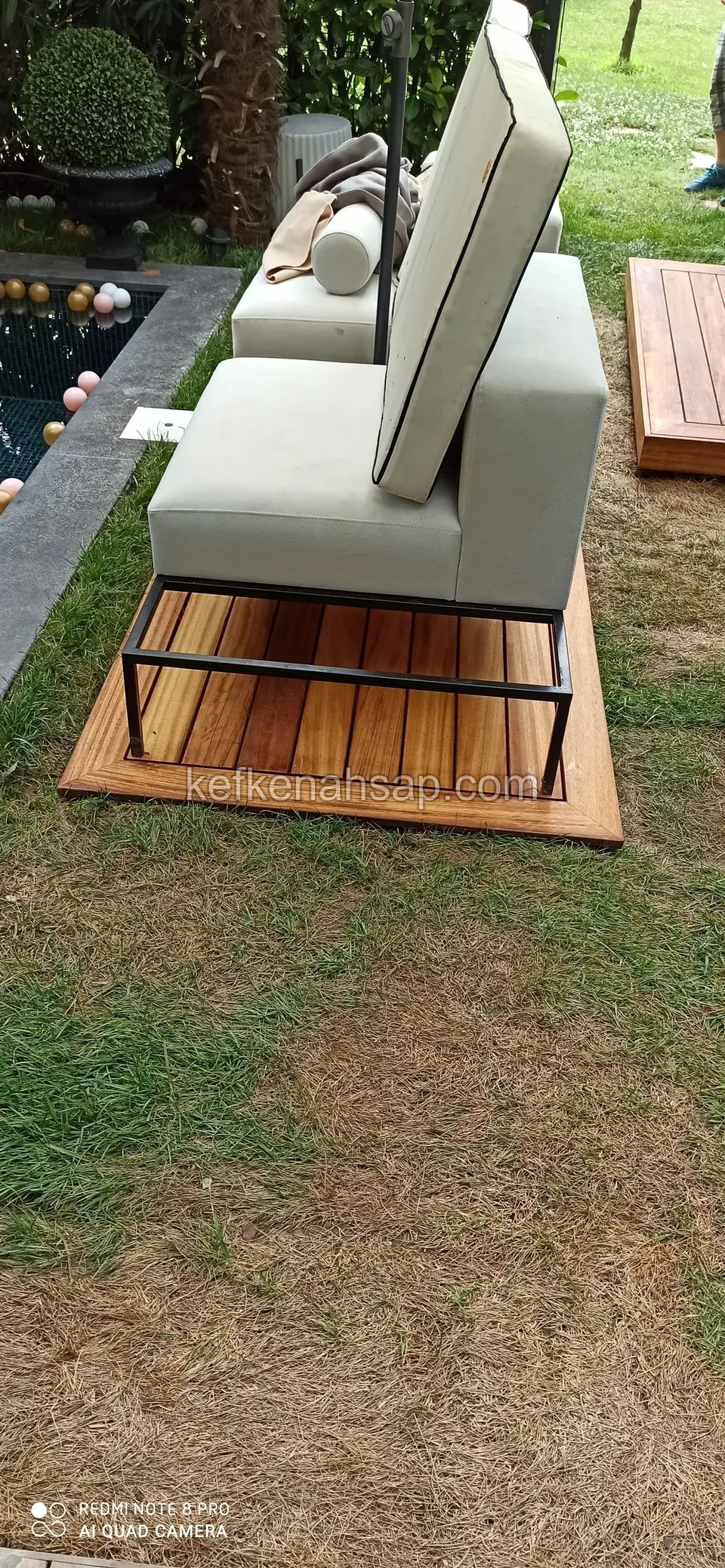 Auf einem Gartenrasen steht ein Sessel mit Metallrahmen und hellen Polstern auf einem kleinen Holzdeckstück
