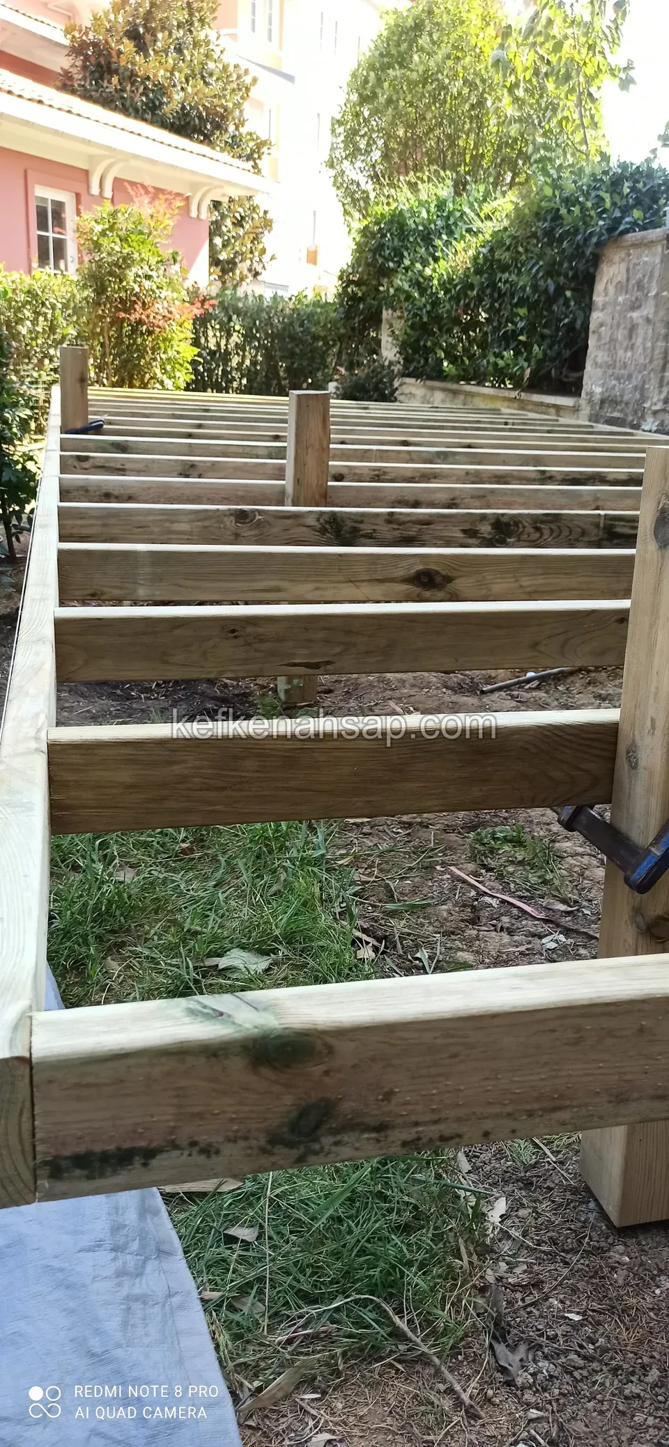 Dicke Holz-Tragbalken über dem Gras für ein aufgeständertes Deck, im Hintergrund ein rosa Gebäude