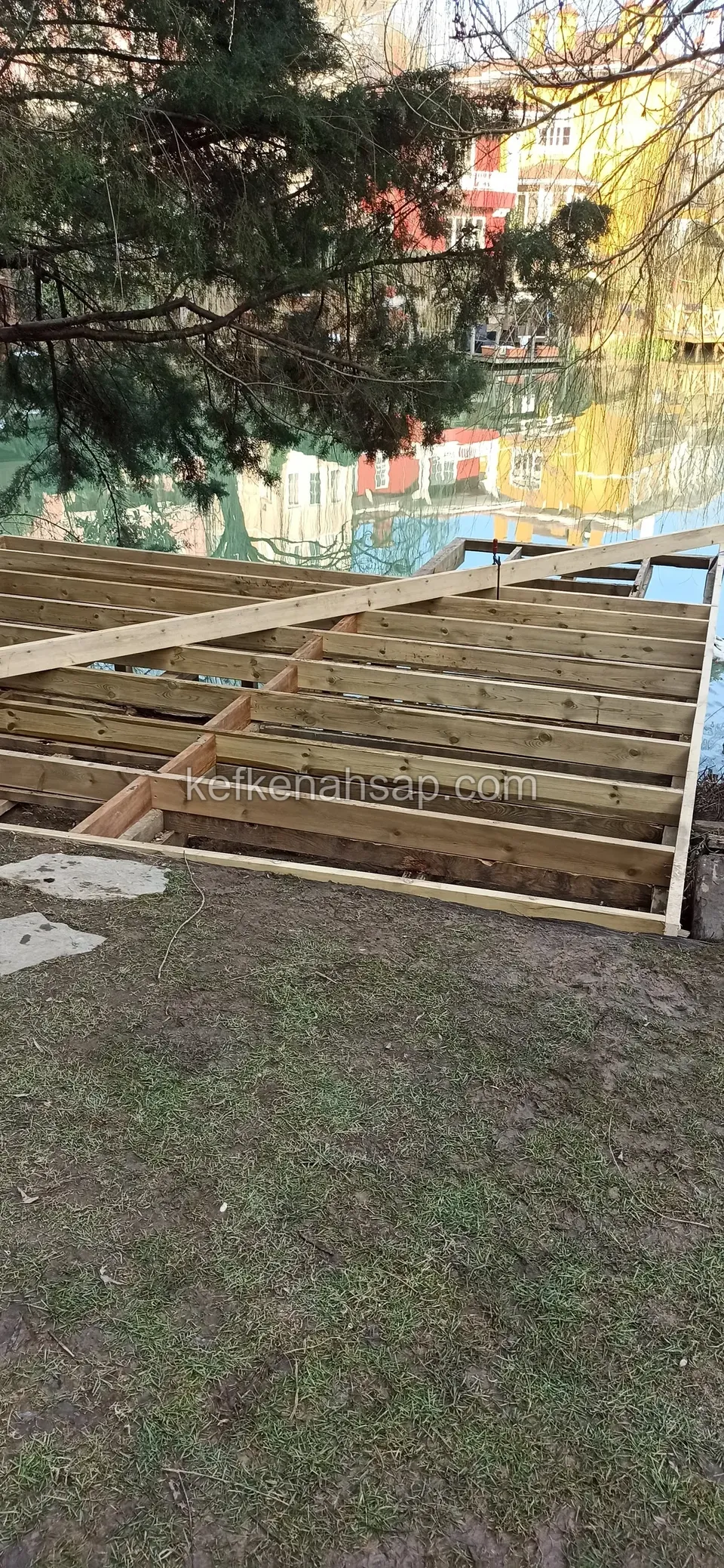 Светлые лаги настила за газоном и ступенчатыми плитами, позади деревья и вода