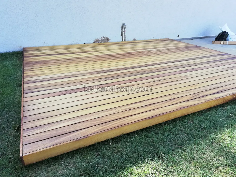 Rötliche Holzdeckplattform aus mehrfach gestreiften Dielen im Rasen vor einer weißen Wand