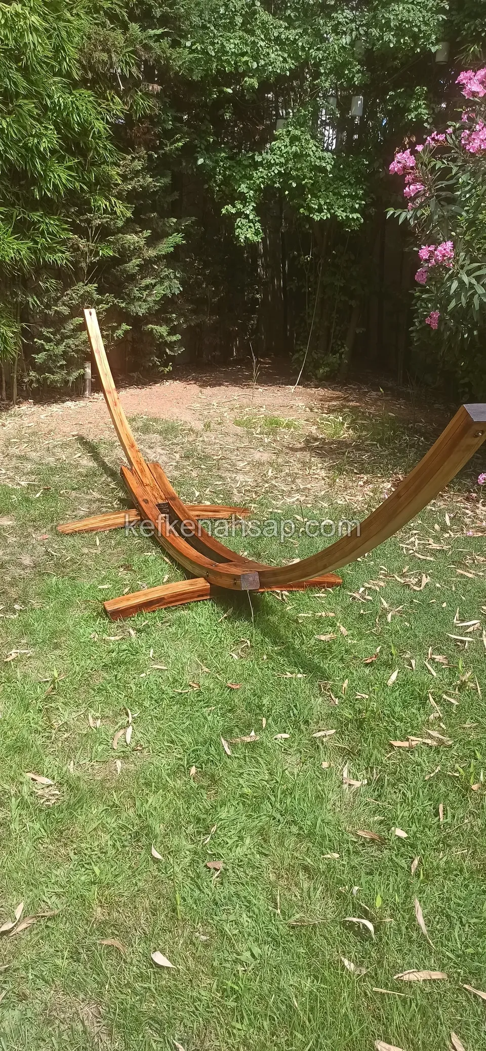 On a garden lawn stands a curved timber hammock stand without its hammock, with bamboo and a pink-flowering tree behind