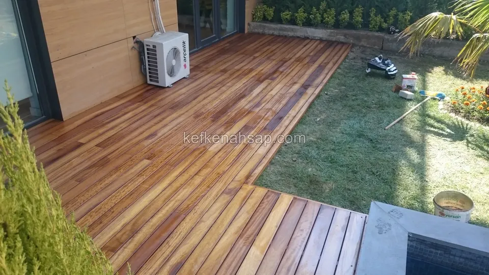 L-shaped deck area outside a house, bordered by lawn on one side, with an air-conditioner unit and greenery beside it
