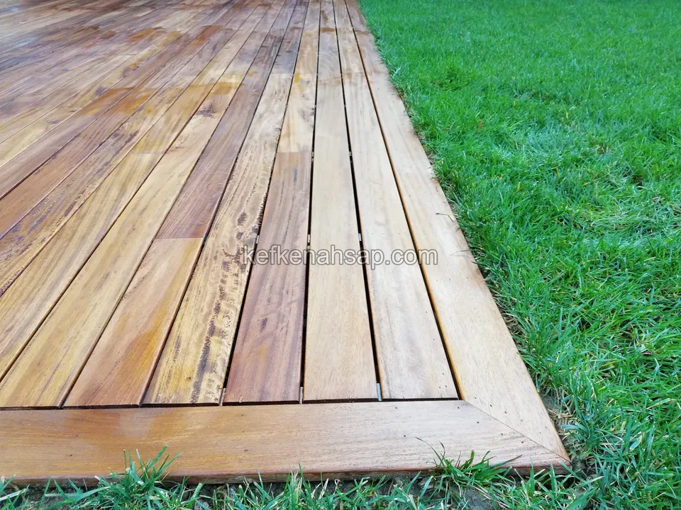 Edge and corner finish of a timber deck beside a lawn, the board surface glossy and red-brown