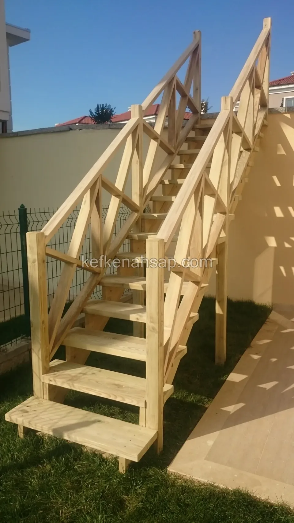 Helle Holztreppe mit Kreuzstreben-Geländern auf beiden Seiten über dem Rasen, dahinter eine Wand und die Dachlinie